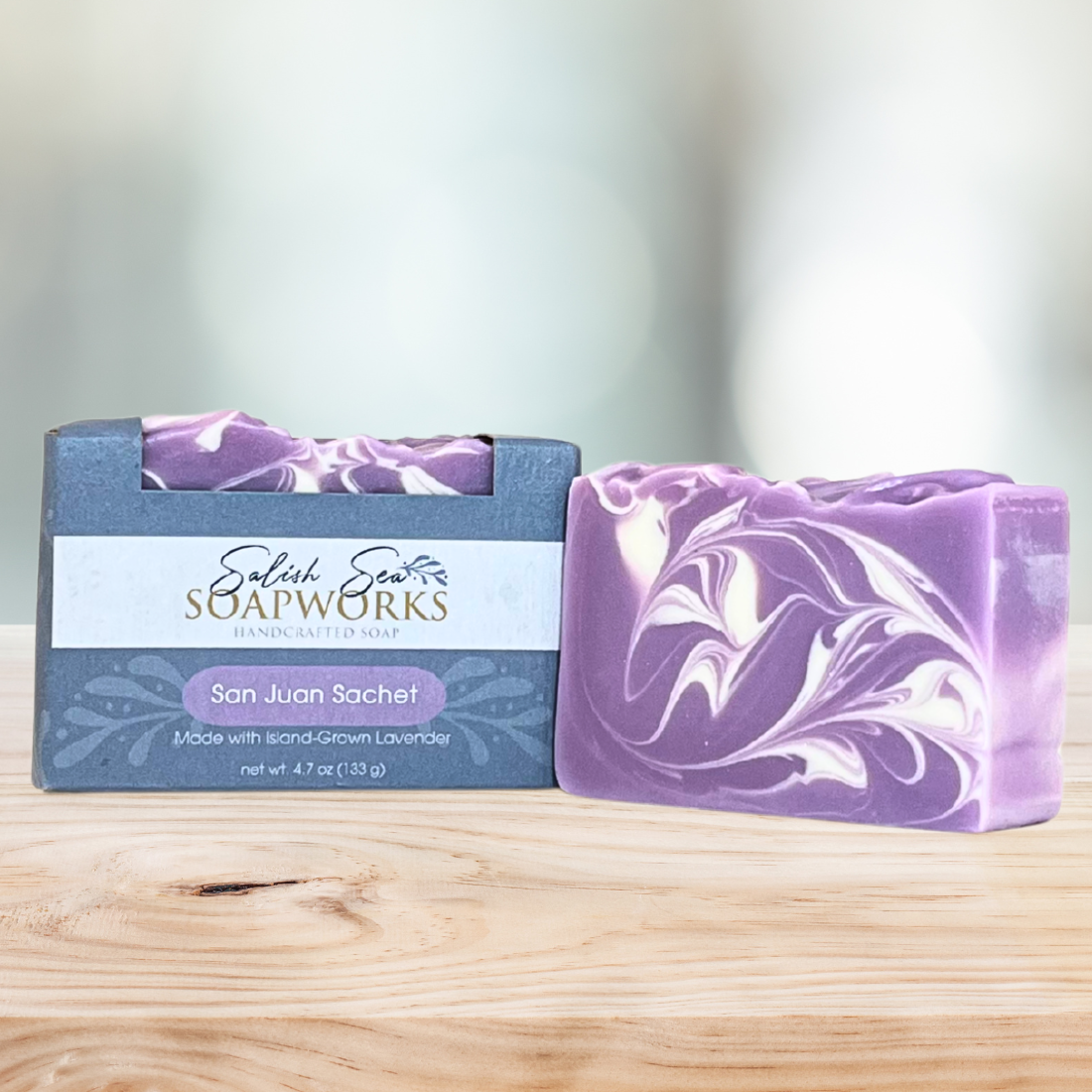 Bar of 'San Juan Sachet' soap in packaging labeled 'Salish Sea Soapworks' on a wooden surface next to a purple and white marbled bar of soap with no packaging.