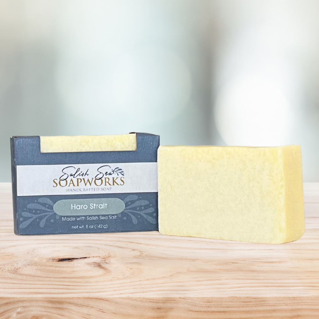 Bar of 'Haro Strait' soap in packaging labeled 'Salish Sea Soapworks' on a wooden surface next to a light yellow bar of soap with no packaging