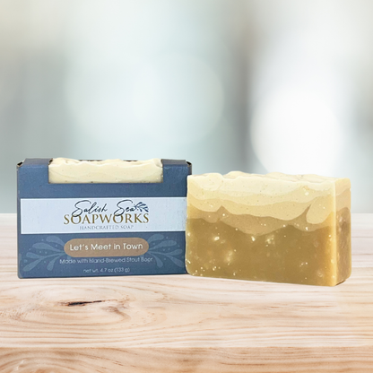 Bar of 'Let's Meet in Town' soap in packaging labeled 'Salish Sea Soapworks' on a wooden surface next to an orange bar of soap with no packaging