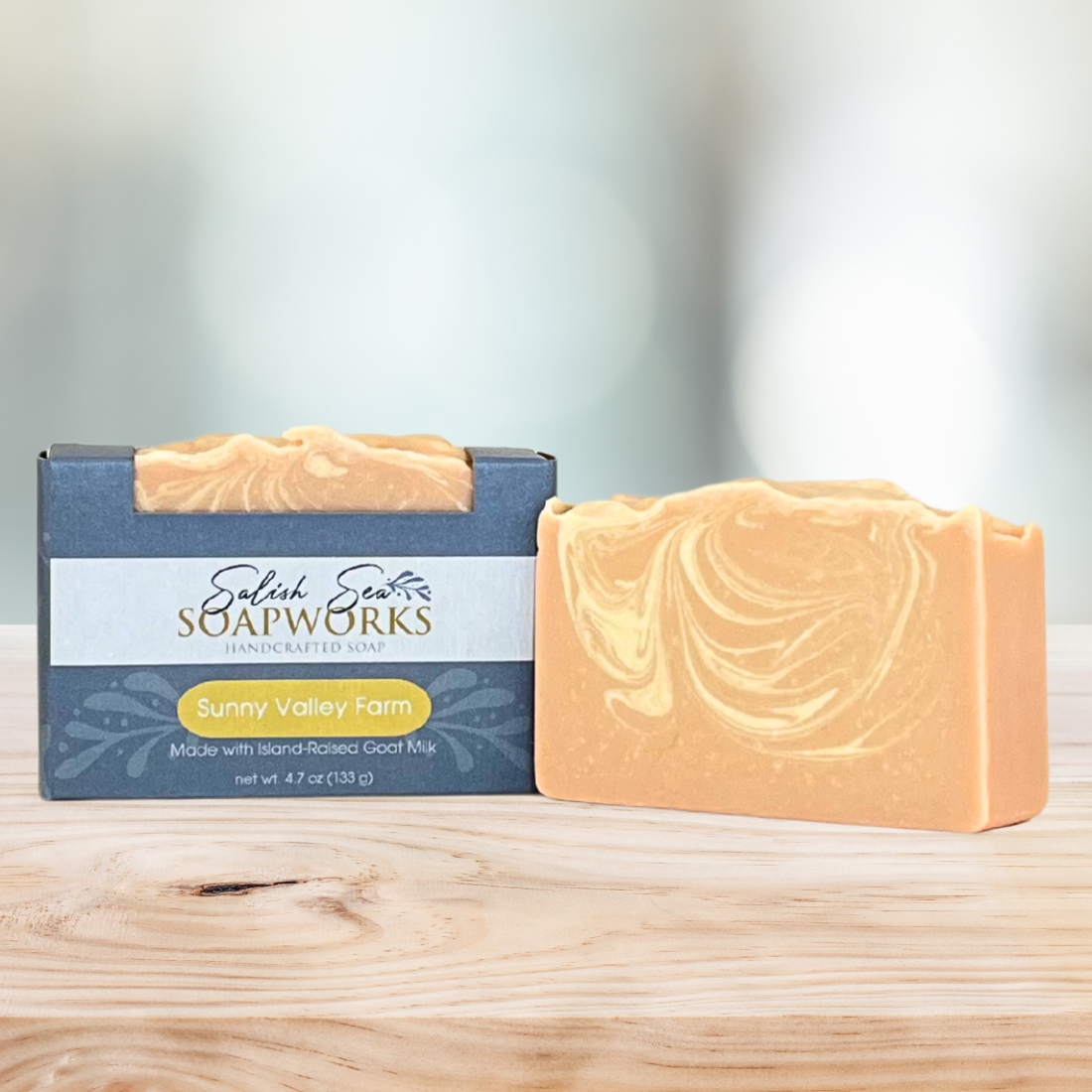 Bar of 'Sunny Valley Farm' soap in packaging labeled 'Salish Sea Soapworks' on a wooden surface next to an orange and yellow marbled bar of soap with no packaging