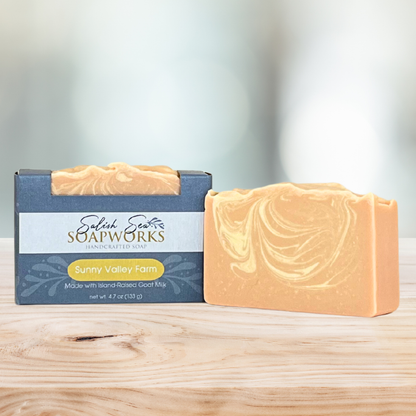 Bar of 'Sunny Valley Farm' soap in packaging labeled 'Salish Sea Soapworks' on a wooden surface next to an orange and yellow marbled bar of soap with no packaging